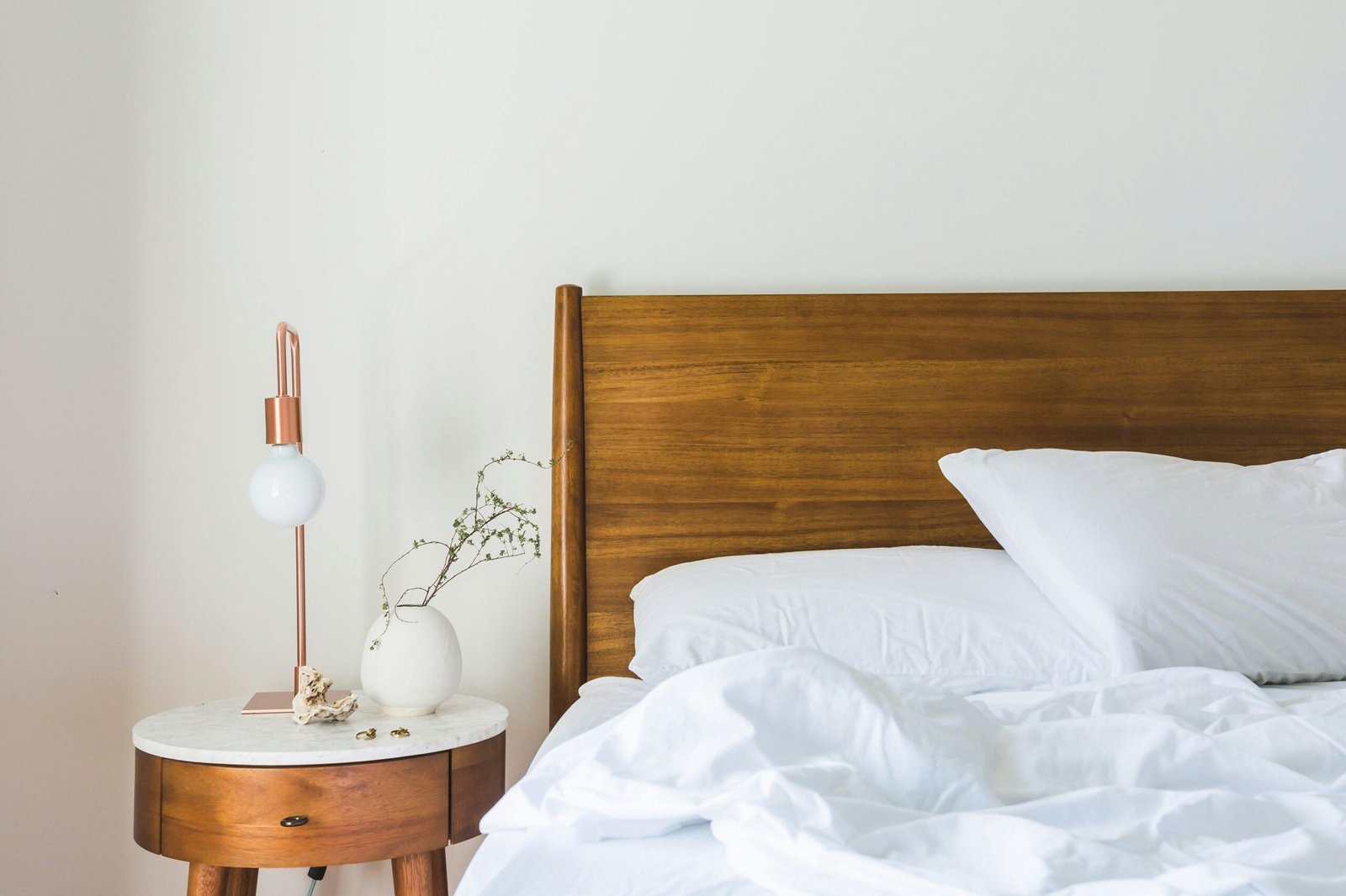 A minimalist bedroom features a wooden headboard, white bedding, and a stylish side table lamp.